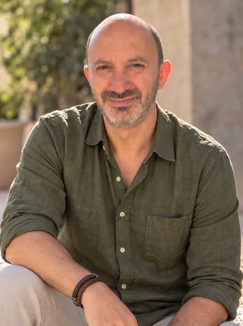 Raul Ortegon Bolio, smiling, in an olive-green shirt, sitting outdoors in the sun with plants and a stone wall behind him.
